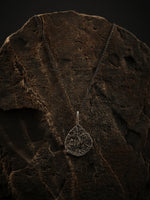 A silver pendant in the shape of a textured teardrop hangs from an oxidized silver chain. The whole rests on a rock.