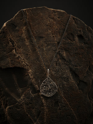 
                  
                    Upload image to gallery, A silver, textured teardrop pendant hangs from an oxidized silver chain. All resting on a rock.
                  
                